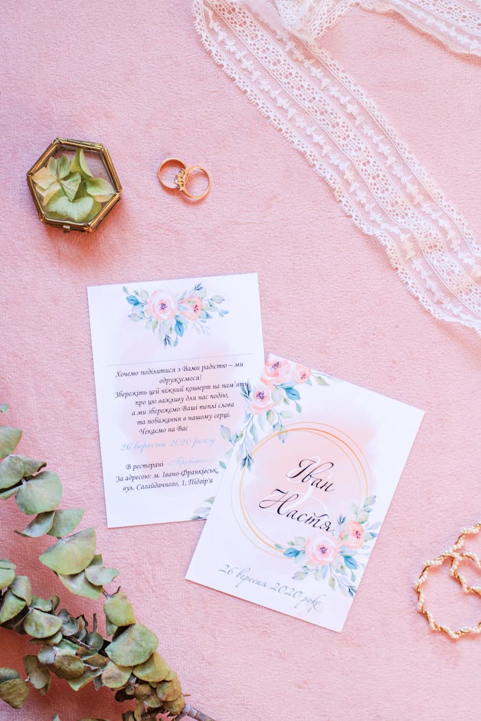 Flat lay of elegant wedding invitations and floral decorations on a pink surface.